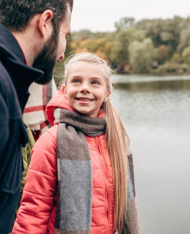 happy father and daughter smiling each other while standing near lake in park